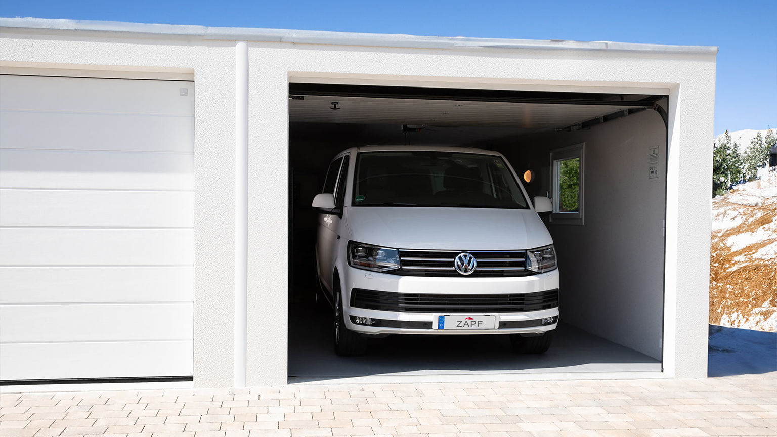 Weißer VW-Bus steht in geöffneter Fertiggarage von ZAPF mit weißem Putz und seitlichem Fenster.