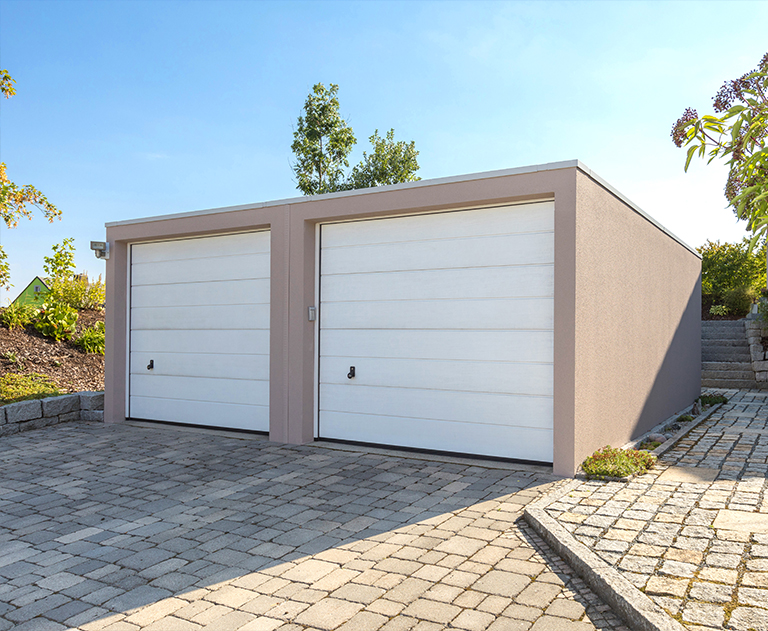 Garage double avec porte blanche et enduit coloré sur une surface pavée en pente.