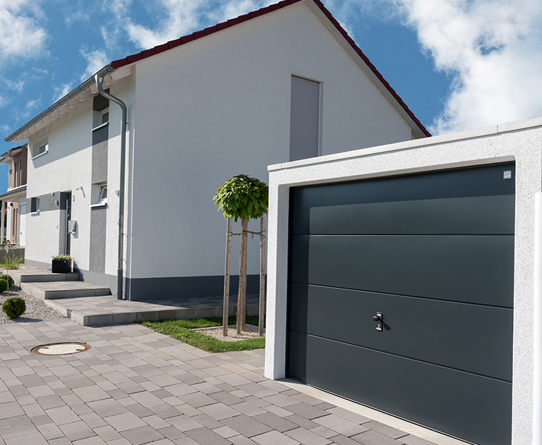 Kompakte Fertiggarage mit anthrazitfarbenem Garagentor vor weißem Einfamilienhaus bei Sonnenschein.
