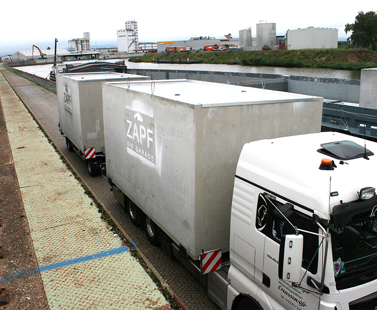 Zwei Schwerlast-Lkw mit aufgeladenen Fertiggaragen von ZAPF auf Hafengelände beim Beladen.