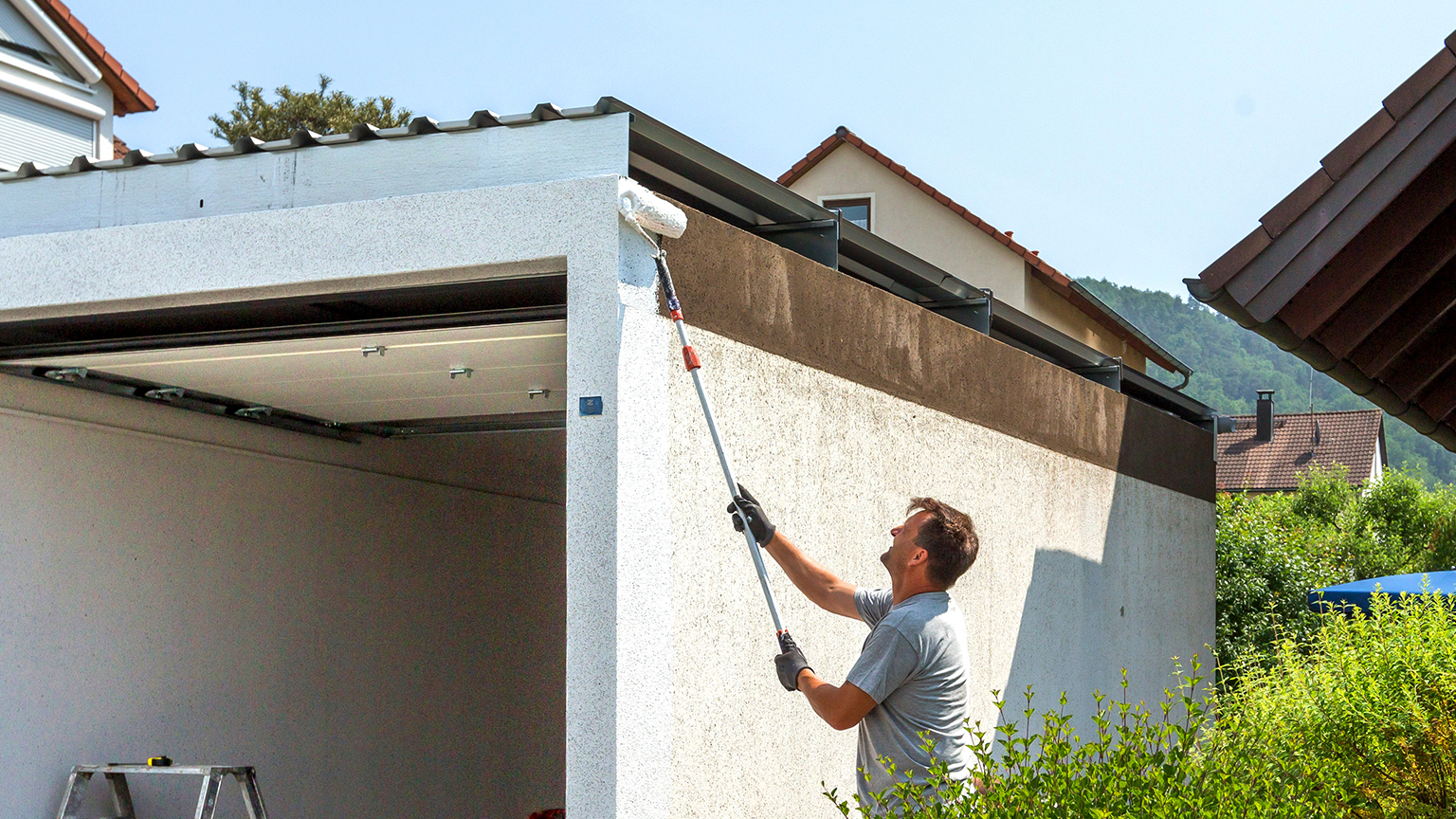 Mann renoviert Garagenfassade, indem er frische weiße Farbe mit einer Rolle aufträgt.