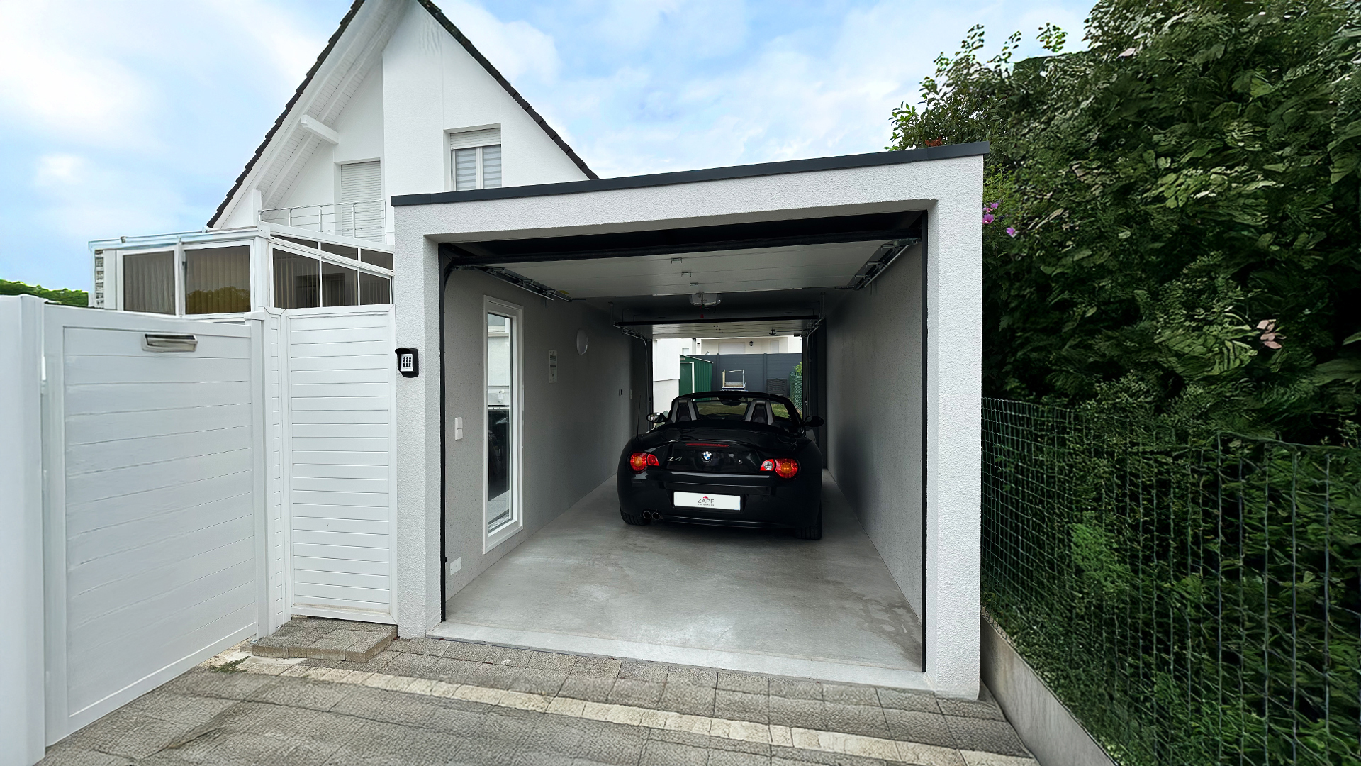 Moderne Einzelgarage mit geöffnetem Tor, in der ein schwarzer BMW Z4 steht. Die Garage ist in ein gepflegtes Wohnumfeld mit weißem Haus, Gartenzaun und Gartentor eingebettet – ein Beispiel für eine genehmigte Garage direkt am Wohnhaus.
