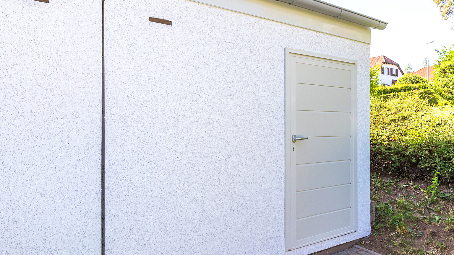 Seitenansicht einer weißen Betonfertiggarage mit Nebeneingangstür, moderner Türfüllung und umliegendem Grün im Hintergrund.