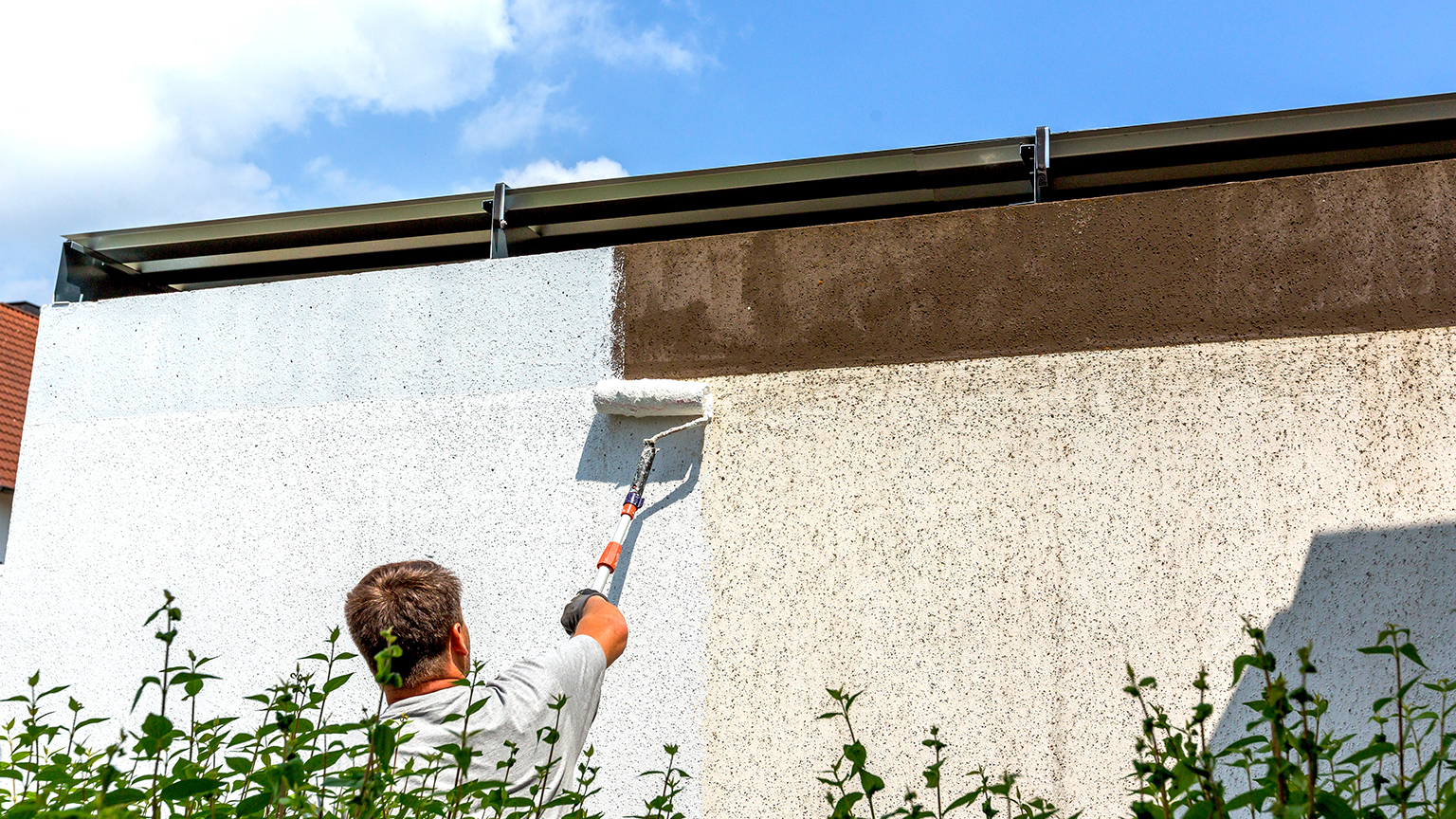 Mann bemalt Außenwand einer Garage mit weißer Farbe und Teleskoprolle.