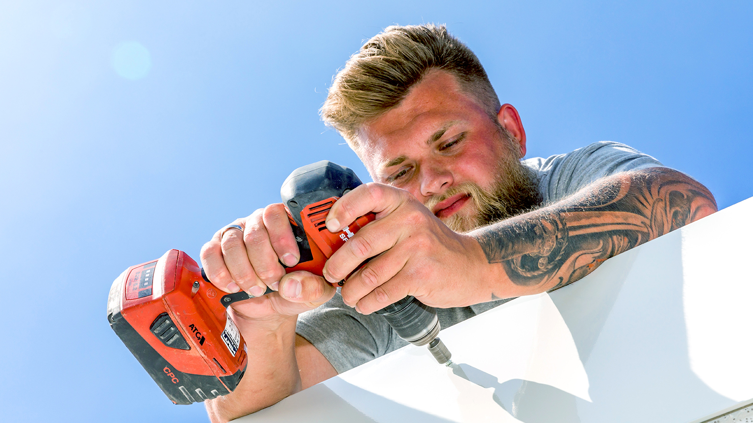 Handwerker verschraubt Metallteil mit Akkuschrauber unter blauem Himmel.