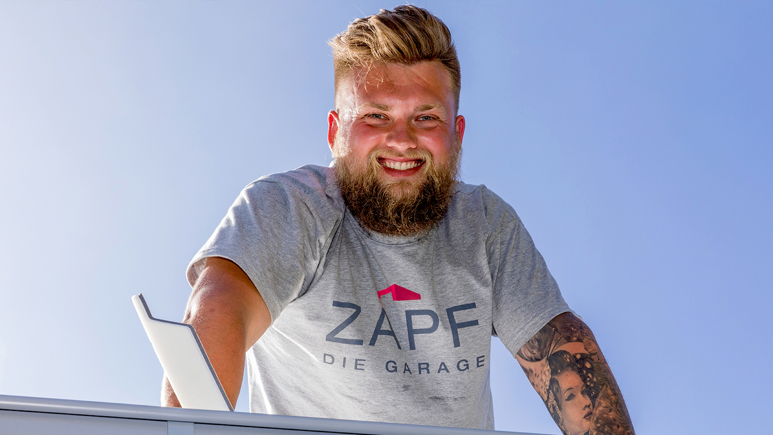 Lachender Mann mit Bart und tätowiertem Arm steht auf einer Garage und trägt ein graues ZAPF T-Shirt.