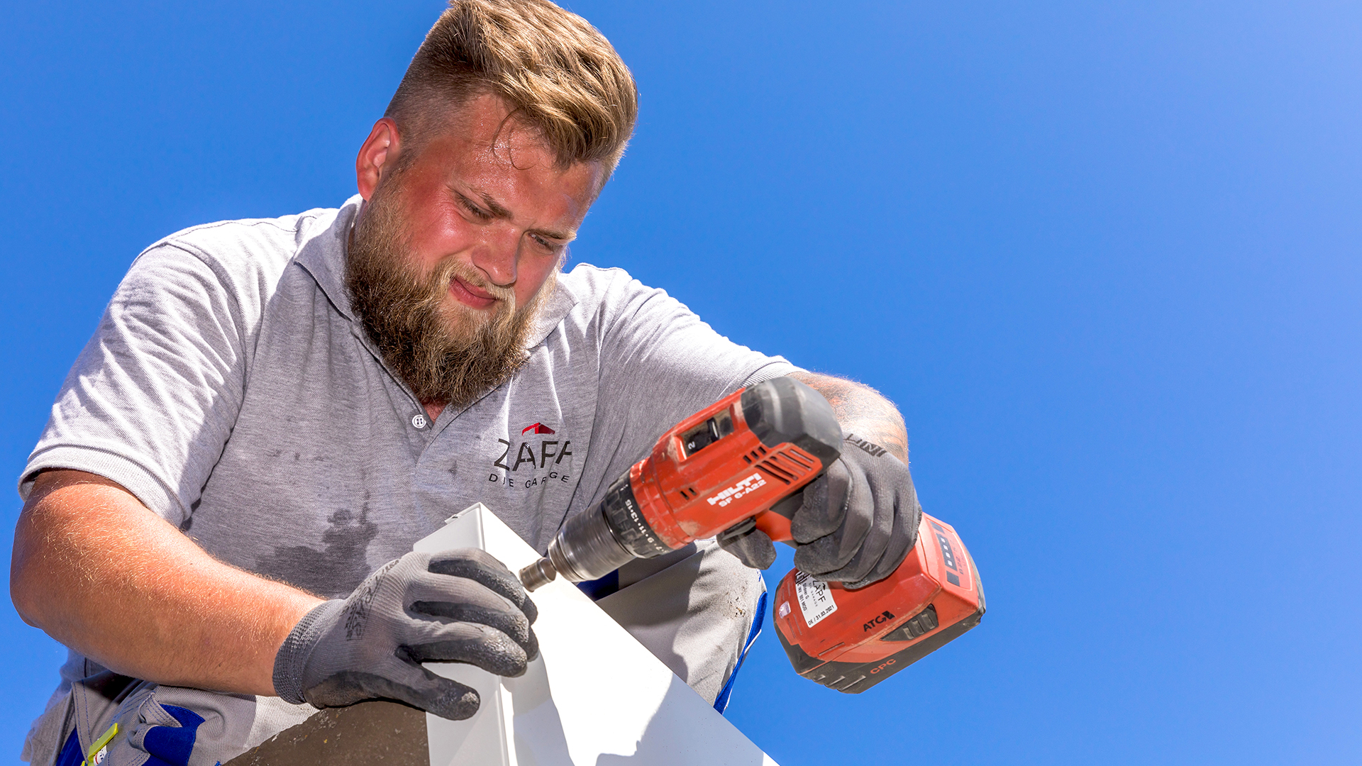 Ein Monteur der Firma ZAPF in grauem Firmenpoloshirt arbeitet konzentriert bei strahlend blauem Himmel auf dem Garagendach. Mit Handschuhen und einem Akkuschrauber befestigt er ein weißes Bauteil – sichtbarer Einsatz und Handwerkskompetenz beim Aufbau einer Fertiggarage.