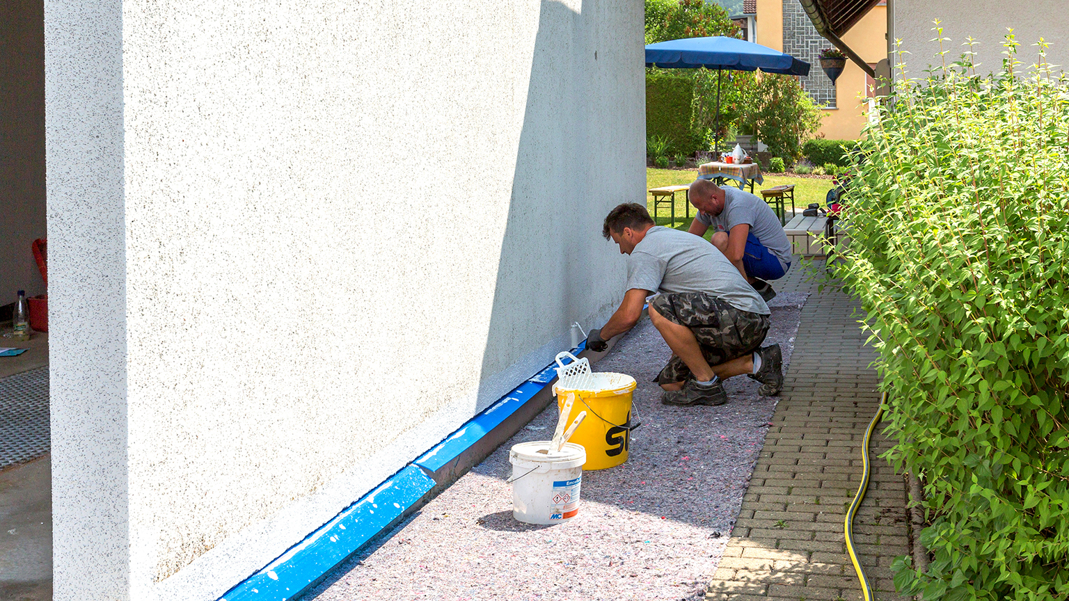 zapf_blog_beitragstitel_punkte Zwei Handwerker streichen den Sockel einer Garage mit weißer Farbe.
