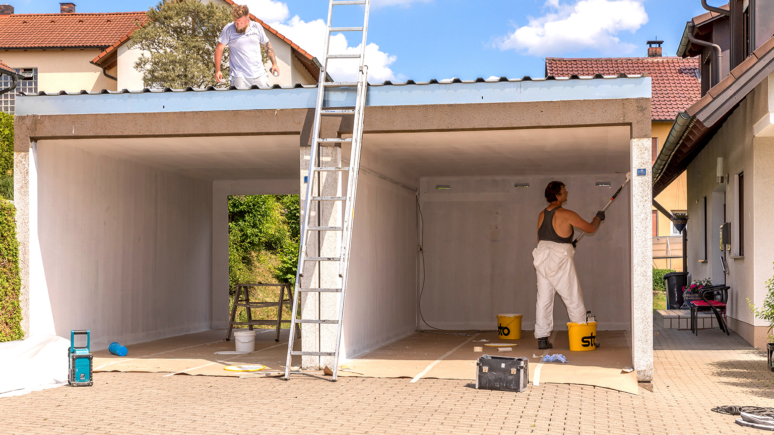 Zwei Handwerker renovieren Doppelgarage, einer arbeitet auf dem Dach, der andere streicht Innenwände.