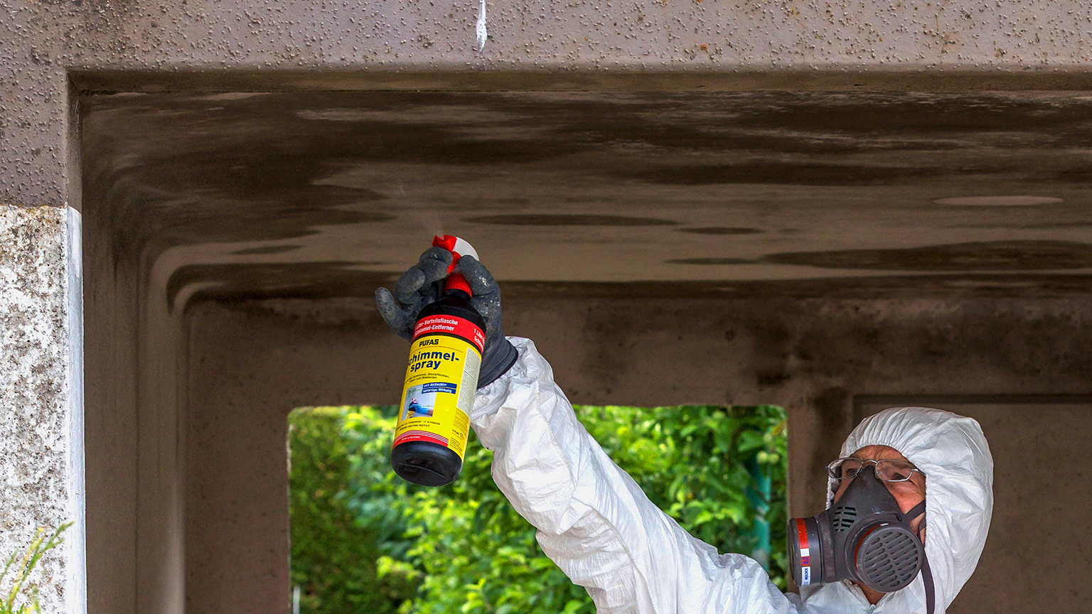 Arbeiter in Schutzanzug und Atemmaske sprüht Schimmelentferner an Garagendecke.