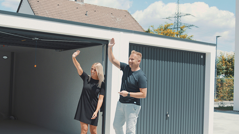 Ein lächelndes Paar steht vor einer modernen Betonfertiggarage mit geöffnetem Garagentor und winkt freundlich – Symbol für Freude über die abgeschlossene Montage oder erteilte Baugenehmigung. Im Hintergrund ein Wohnhaus und blauer Himmel mit vereinzelten Wolken.