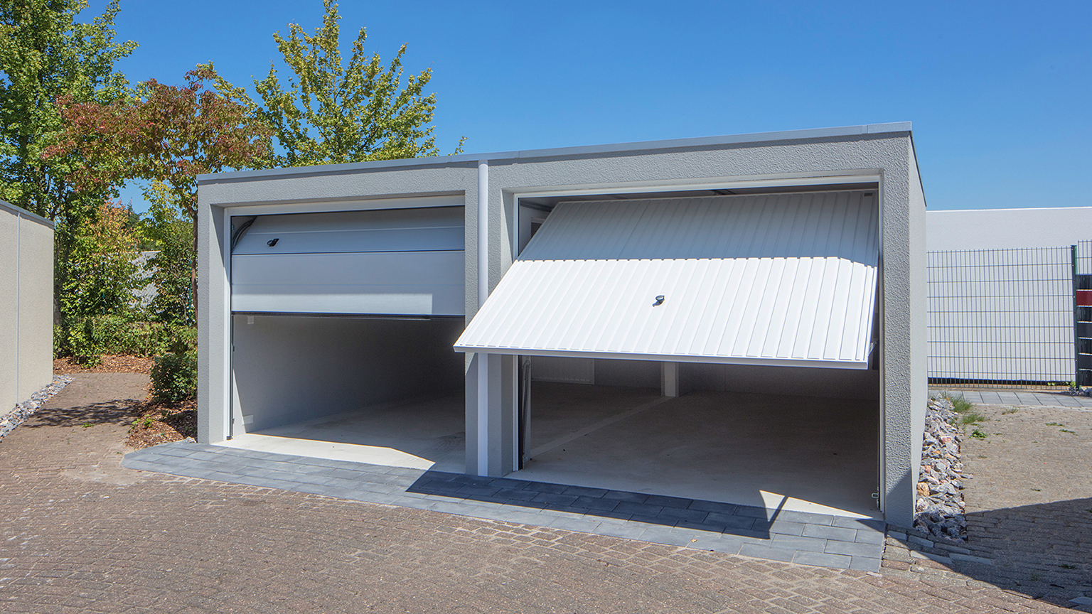 zapf_blog_beitragstitel_handwerker_1 Zwei moderne Betonfertiggaragen mit geöffneten Schwingtoren, nebeneinander aufgestellt. Die Garagen haben eine helle, glatte Fassade und stehen auf einem gepflasterten Hof. Eine Garage hat das Tor halb geöffnet, die andere vollständig hochgeklappt. Im Hintergrund sind grüne Bäume und ein klarer blauer Himmel zu sehen.