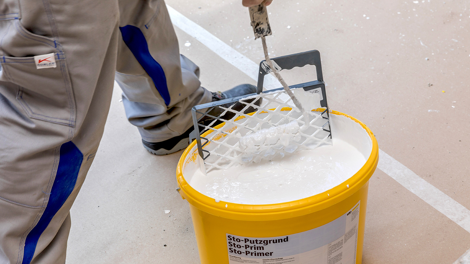 zapf_blog_beitragstitel_handwerker_3 Handwerker taucht eine Farbrolle in einen Eimer mit weißem Putzgrund. Der gelbe Eimer ist mit dem Etikett „Sto-Putzgrund Sto-Prim Sto-Primer“ versehen. Der Boden ist mit Malervlies abgedeckt, und der Arbeiter trägt graue Arbeitshose mit blauen Einsätzen.