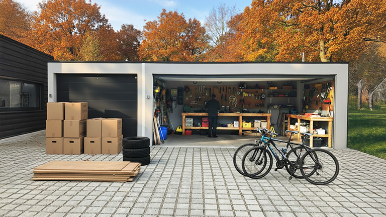 Großzügige ZAPF Garage mit geöffneter Tür, eingerichtet als Heimwerkstatt. Eine Person arbeitet an einer Werkbank, umgeben von Werkzeugen und Regalen. Vor der Garage stehen Fahrräder, Kartons und Autoreifen auf einer gepflasterten Einfahrt, im Hintergrund herbstlich gefärbte Bäume.