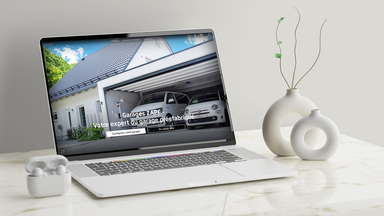 Laptop auf einem hellen Marmortisch mit geöffneter Website von ZAPF Garagen. Auf dem Bildschirm ist eine moderne Doppelgarage mit einem weißen Auto zu sehen. Text auf dem Display: „Garages ZAPF – Votre expert du garage préfabriqué“. Die neue Website zeigt, dass ZAPF Garagen nun auch in Englisch und Französisch verfügbar ist.