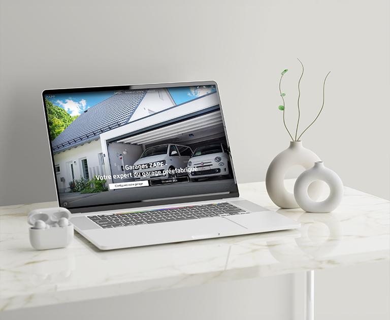 Ordinateur portable sur une table en marbre affichant le nouveau site web de ZAPF Garages. On y voit un garage double moderne avec une voiture blanche. Texte à l’écran : « Garages ZAPF – Votre expert du garage préfabriqué ». Le site est désormais disponible en français et en anglais.