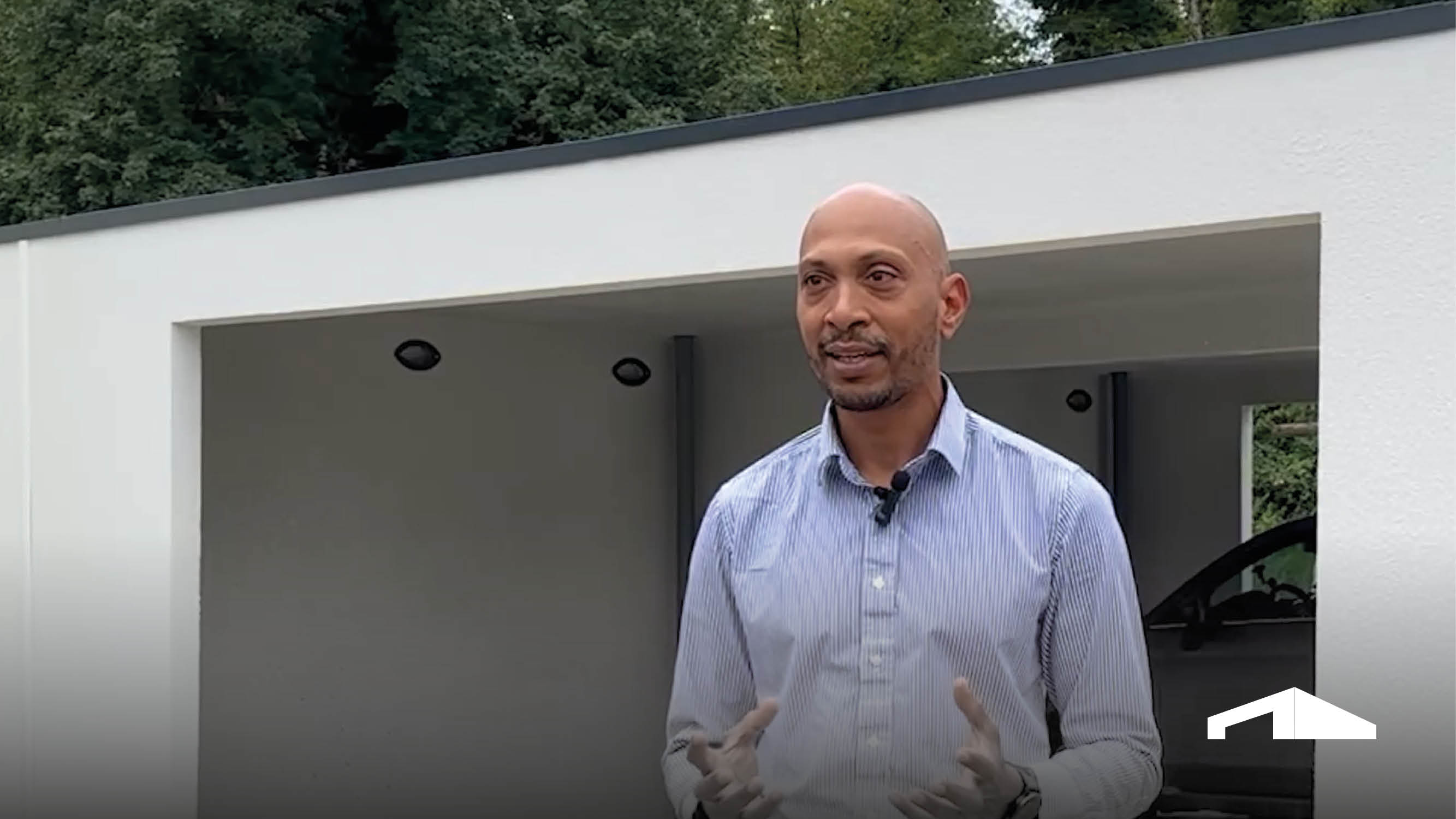 Mann steht vor moderner weißer Doppelgarage mit Carport-Funktion und spricht, Fahrzeug im Hintergrund sichtbar.