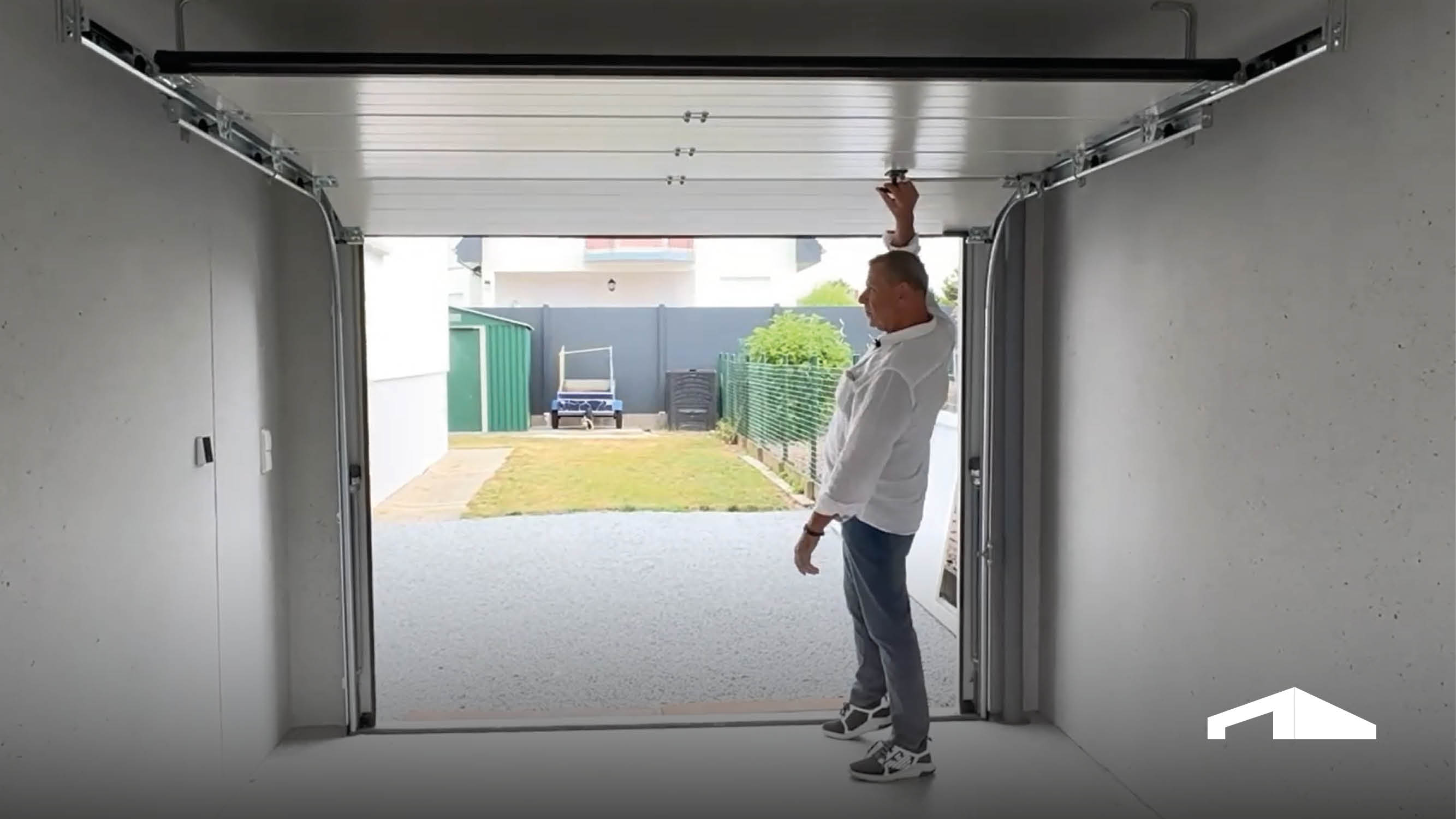 Mann steht in moderner Garage und öffnet das hintere Sektionaltor per Hand, Blickrichtung nach außen auf Garten und Einfahrt.