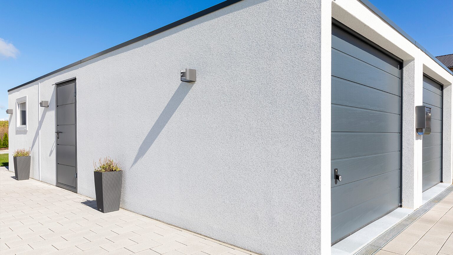 Doppelgarage mit Anbau Moderne Betonfertig-Doppelgarage mit Anbau, heller Putzfassade und Flachdach. Rechts zwei graue Sektionaltore der Doppelgarage, links ein seitlicher Anbau mit Eingangstür, Fenster und Außenleuchten. Die Garage steht auf gepflasterter Fläche in moderner Wohnumgebung, von ZAPF Garagen.