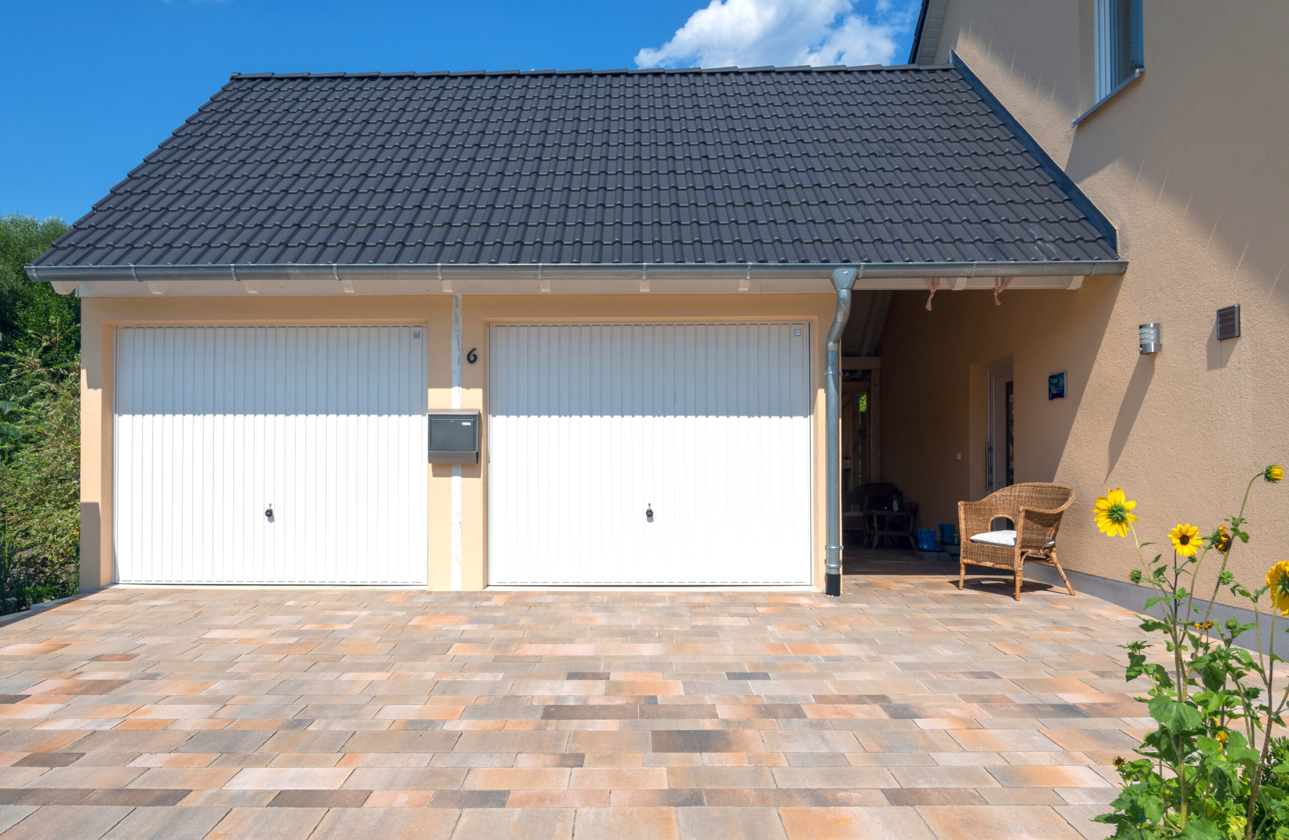 ZAPF-Doppelgarage mit Dachaufbau Doppelgarage mit Satteldach und zwei weißen Garagentoren, integriert in ein Wohngebäude. Die Garage verfügt über eine helle Putzfassade, gepflasterte Einfahrt und ein geneigtes Ziegeldach. Rechts ist ein überdachter Durchgang zum Haus erkennbar, von ZAPF Garagen.