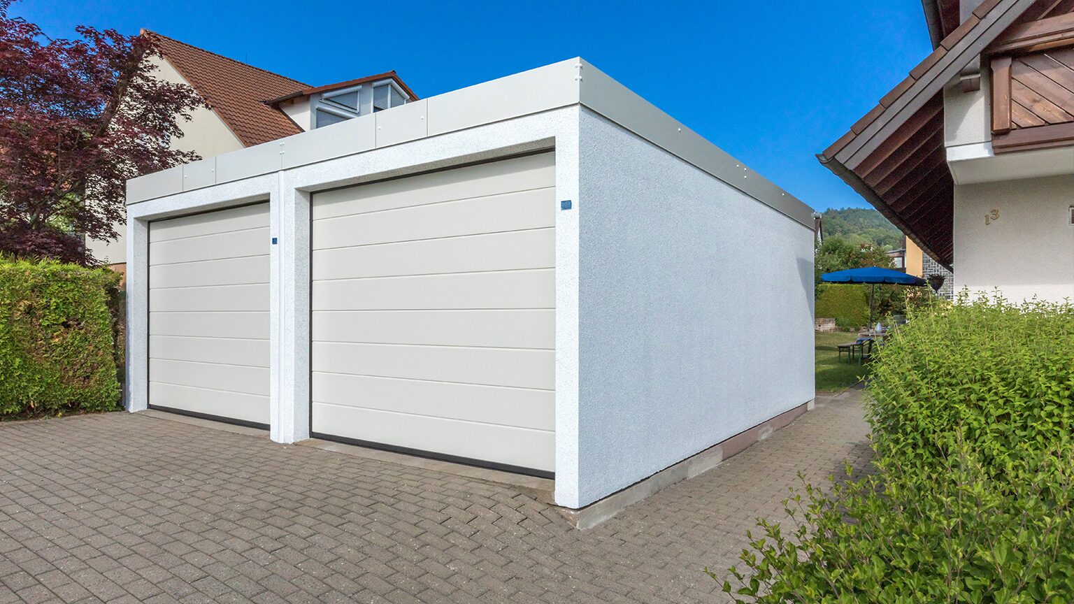 ZAPF Pultdach Moderne ZAPF Doppel-Fertiggarage aus Beton mit hellem Putz, zwei weißen Sektionaltoren und flachem Pultdach in klarer, kubischer Bauform, freistehend im Wohnumfeld, von ZAPF.