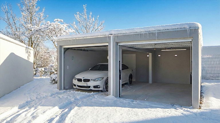 Moderne Betonfertiggarage als Doppelgarage im Winter, mit zwei offenen Garagentoren und schneebedecktem Flachdach. In einer Garage steht ein weißes Auto, die zweite Stellfläche ist leer. Der Innenraum wirkt hell, mit glatten Betonwänden und integrierter Beleuchtung, umgeben von verschneiter Außenanlage, von ZAPF Garagen.