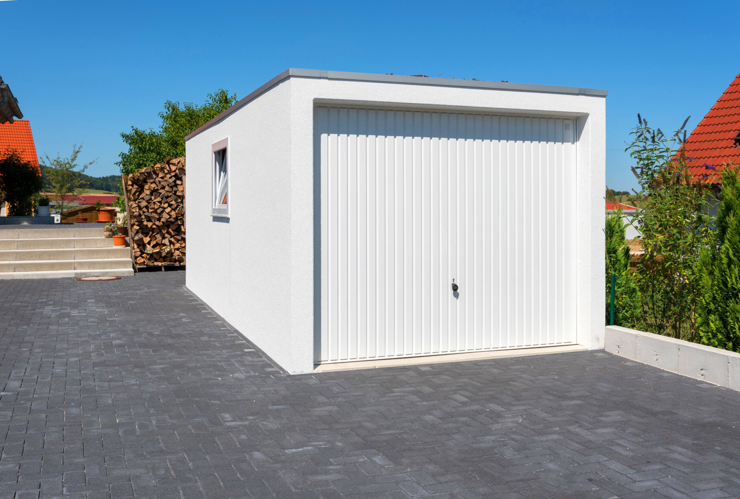 Moderne Einzelgarage mit weißem Schwingtor Freistehende Betonfertiggarage mit weiß verputzter Fassade und vertikal strukturiertem Schwingtor auf gepflasterter Einfahrt, seitlichem Fenster und Flachdach in einem Wohngebiet, ZAPF Garagen