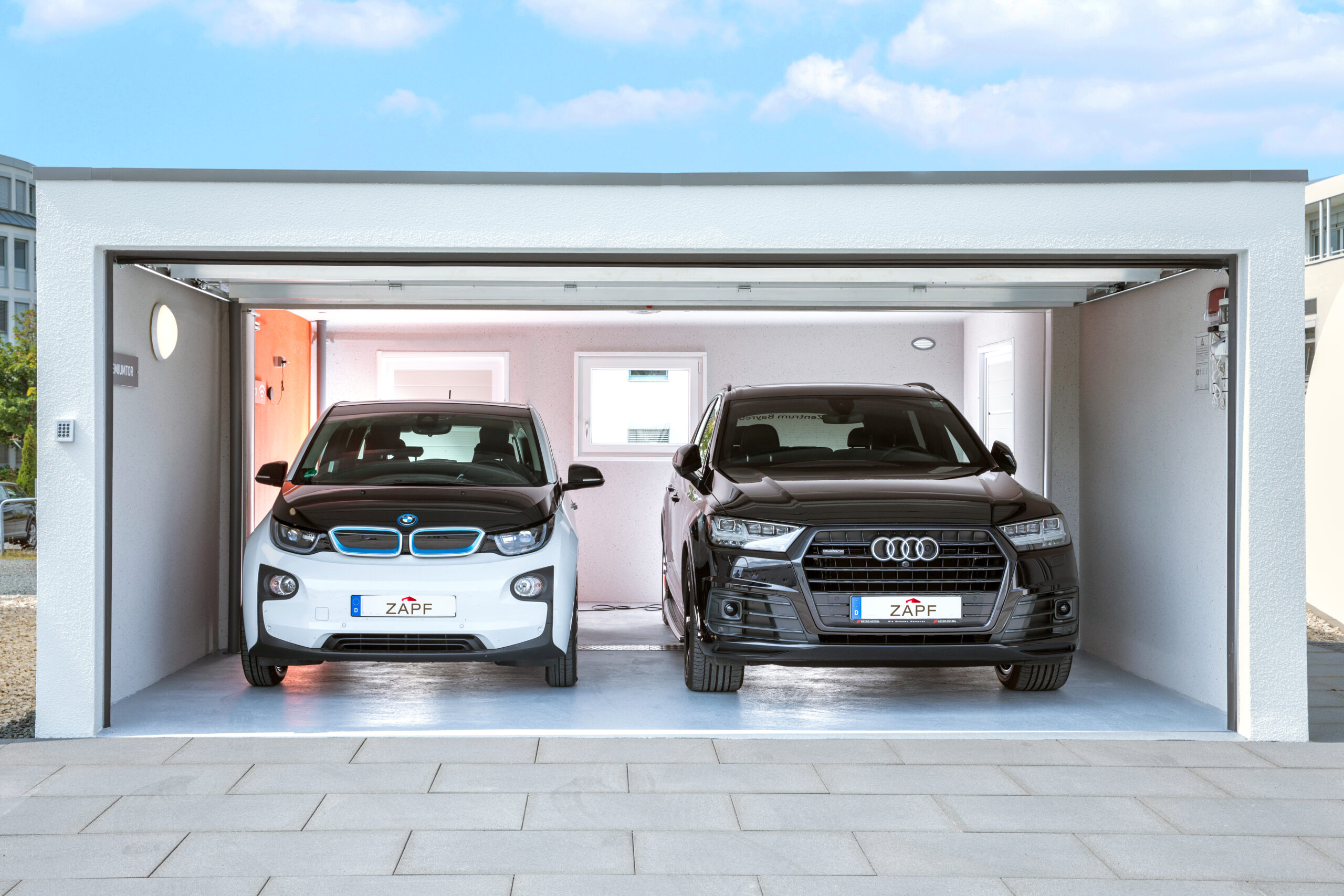 Offene Doppelgarage mit zwei Fahrzeugen Innenansicht einer Betonfertig-Doppelgarage mit geöffnetem Garagentor und zwei geparkten Fahrzeugen, glatten Innenwänden, Fenster und Beleuchtung, auf einer gepflasterten Einfahrt vor modernem Gebäude, ZAPF Garagen