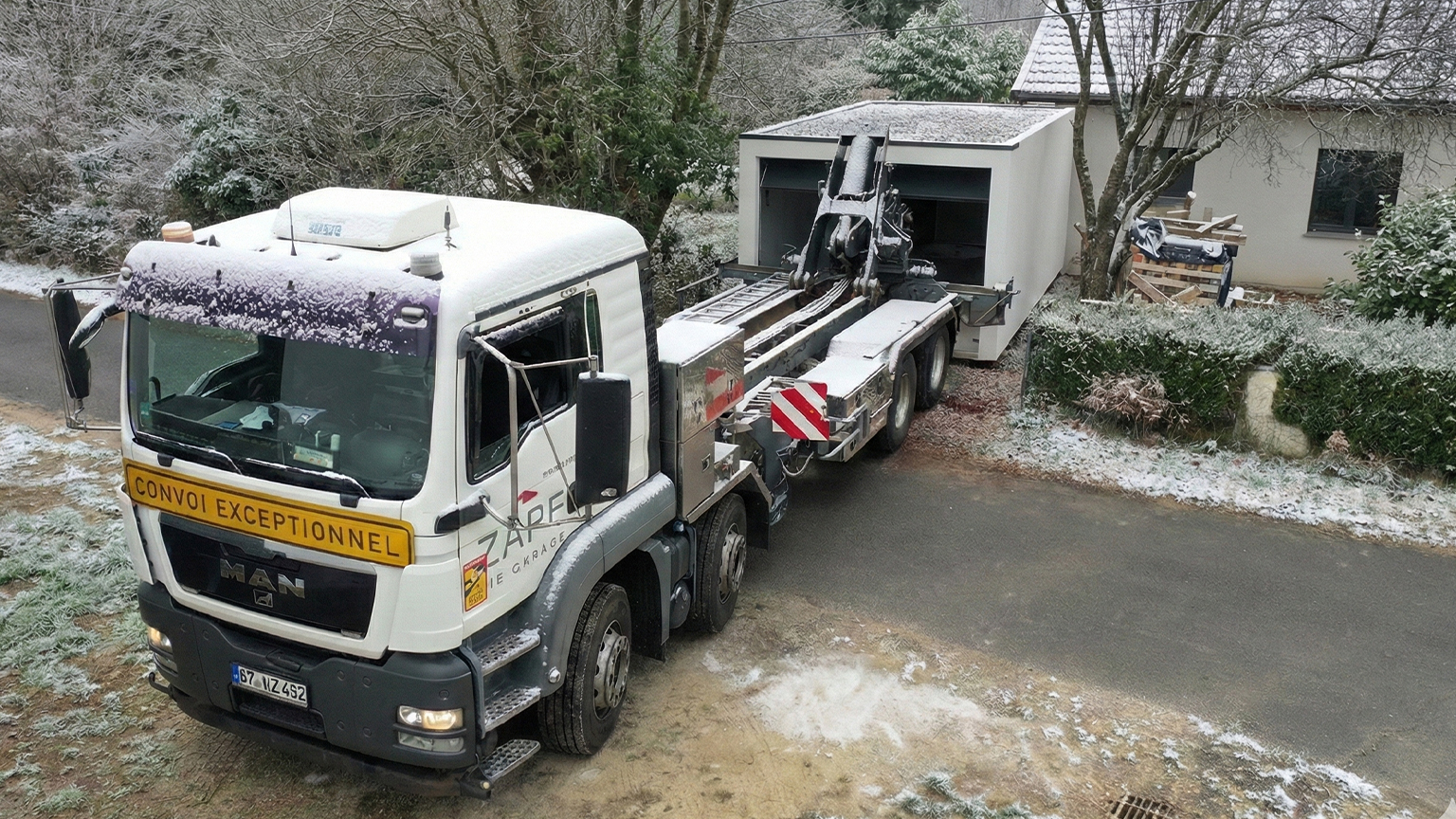 Anlieferung einer Betonfertiggarage mit Spezialfahrzeug im Winter Spezial-Lkw mit Kran bringt eine weiße Betonfertiggarage in eine private Einfahrt bei winterlichen Bedingungen, von ZAPF