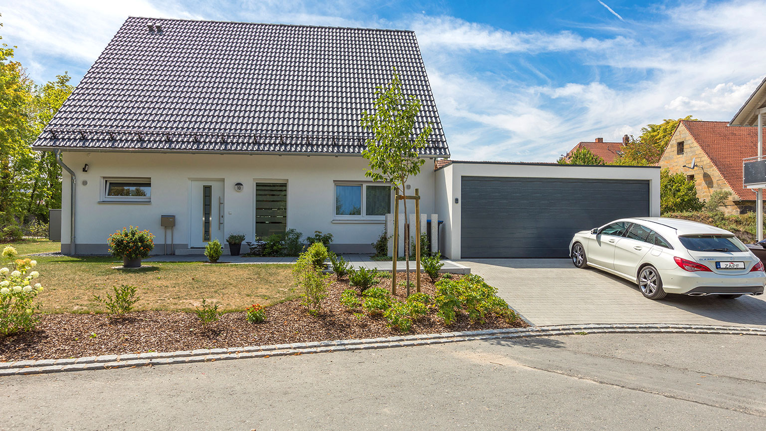 zapf_blog_beitragstitel_einfamilienhaus-satteldach-garage-anthrazit-sektionaltor-kombi Modernes Einfamilienhaus mit Satteldach, angebauter Garage mit anthrazitfarbenem Sektionaltor und weißem Kombi auf gepflasterter Einfahrt.