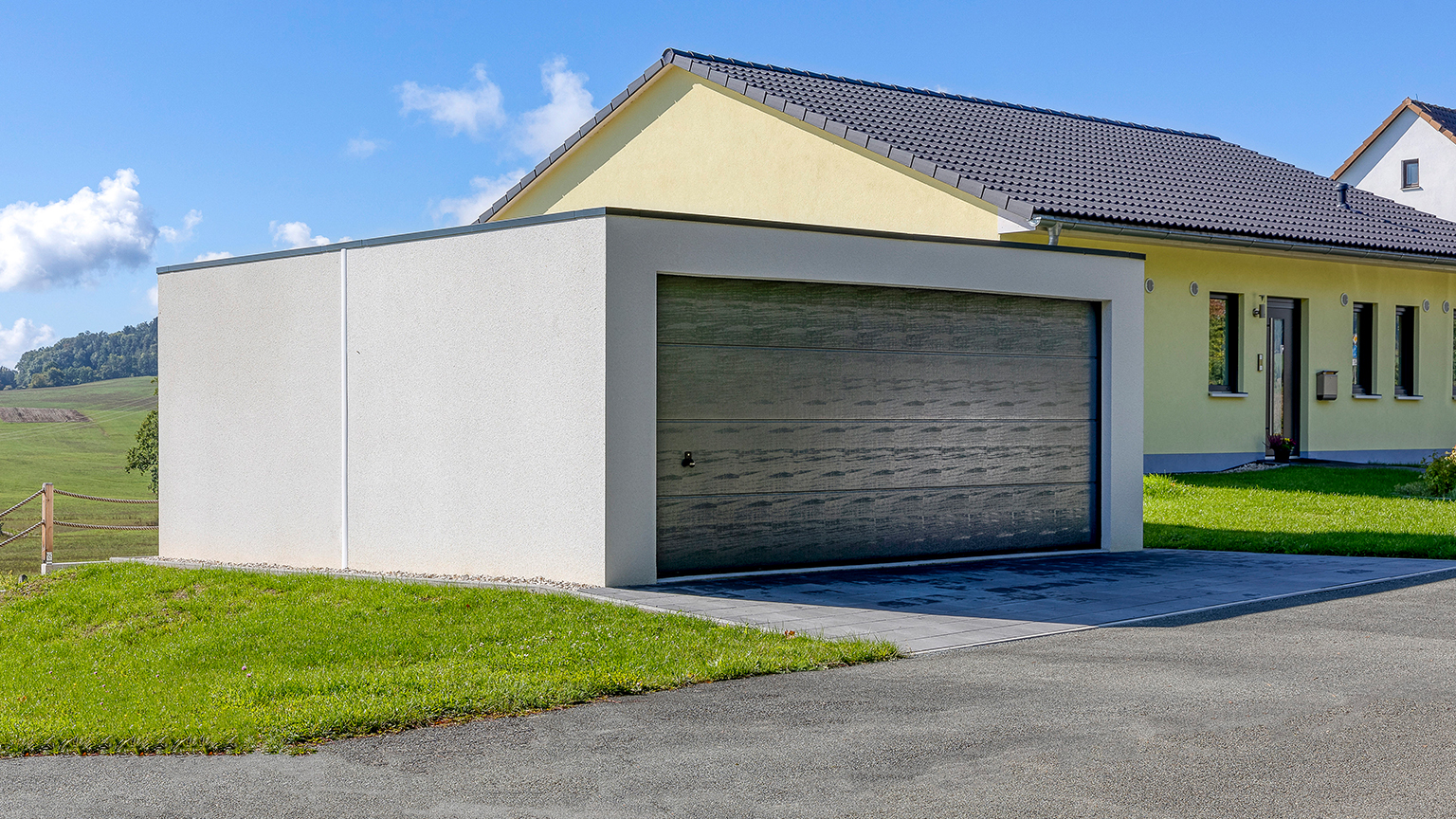 Garage préfabriqué en béton moderne avec large porte sectionnelle à côté d’une maison individuelle Garage préfabriqué en béton indépendant avec toit plat et large porte sectionnelle aspect bois, implanté à côté d’une maison individuelle à toit en pente, entouré d’une pelouse et d’une allée aménagée du fabricant ZAPF