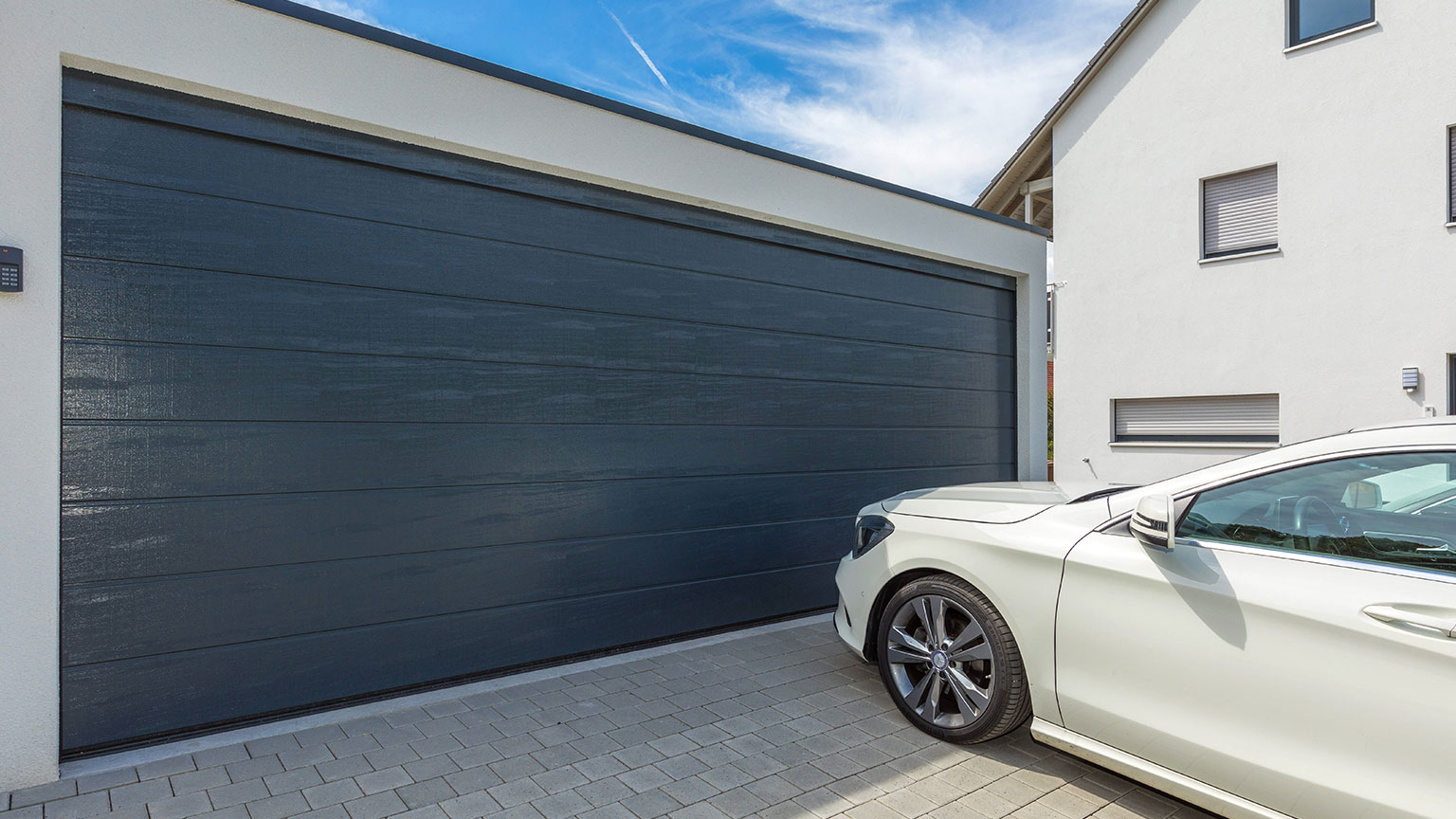 zapf_blog_beitragstitel_flachdachgarage-anthrazit-sektionaltor-weisses-auto-pflastereinfahrt Großes anthrazitfarbenes Sektionaltor an moderner Flachdachgarage mit weißer Fassade, davor ein weißes Auto auf gepflasterter Einfahrt.