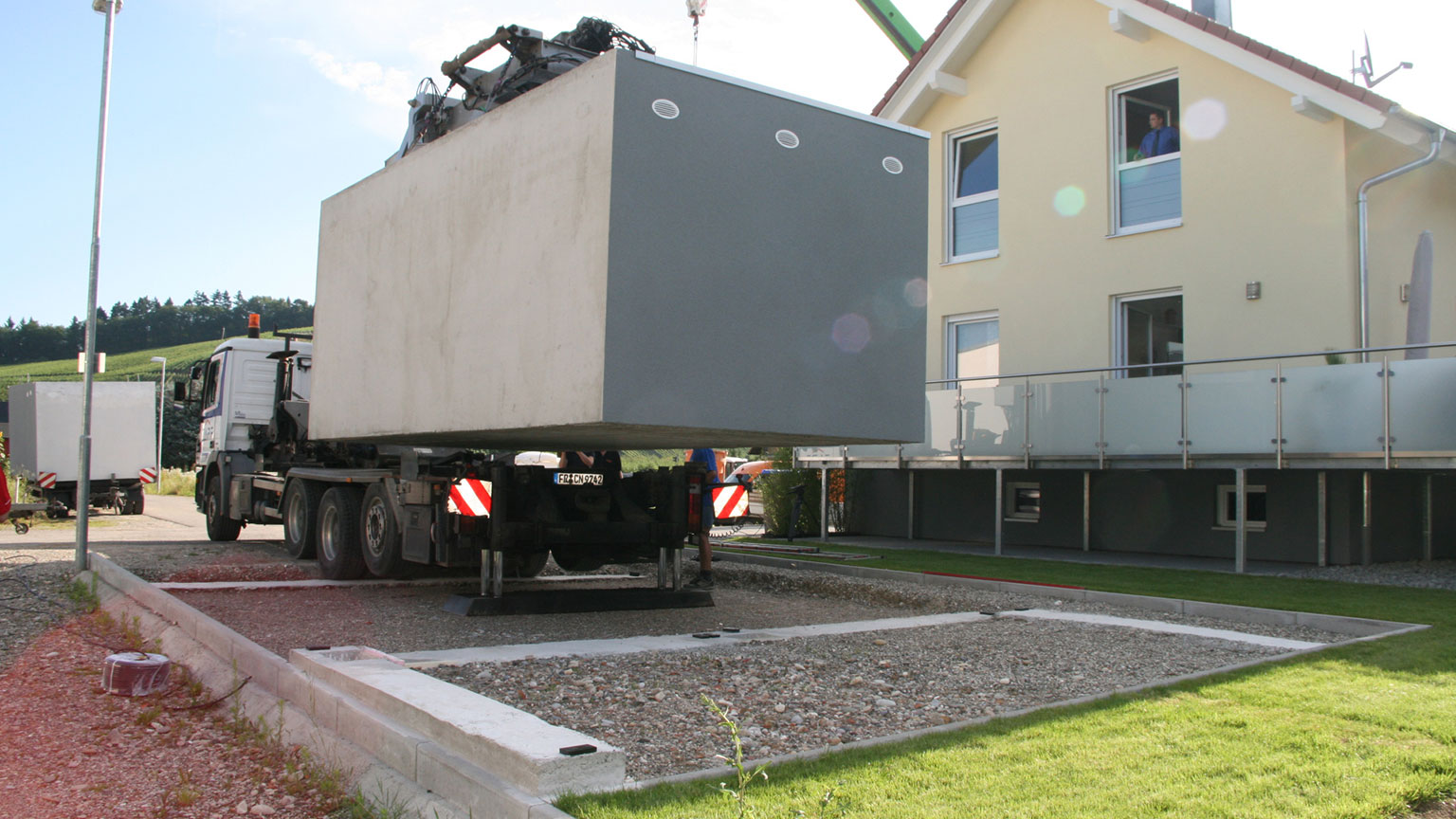 Fertiggarage aus Beton wird per Lkw-Kran versetzt Versetzen einer Betonfertiggarage mit Lkw-Kran auf ein vorbereitetes Streifenfundament neben einem Wohnhaus, Garagenkörper schwebt über der Einbaufläche, ZAPF Qualität vom Hersteller ZAPF