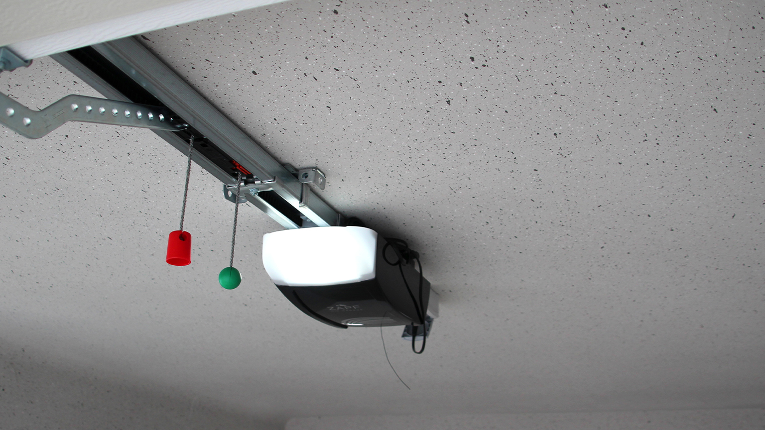 Garagentorantrieb an Betondecke montiert Elektrischer Garagentorantrieb mit Führungsschiene unter einer hellen Betondecke montiert, sichtbare Notentriegelung mit rotem und grünem Zugknauf sowie integrierte Beleuchtungseinheit in einer Betonfertiggarage in ZAPF Qualität von ZAPF Garagen