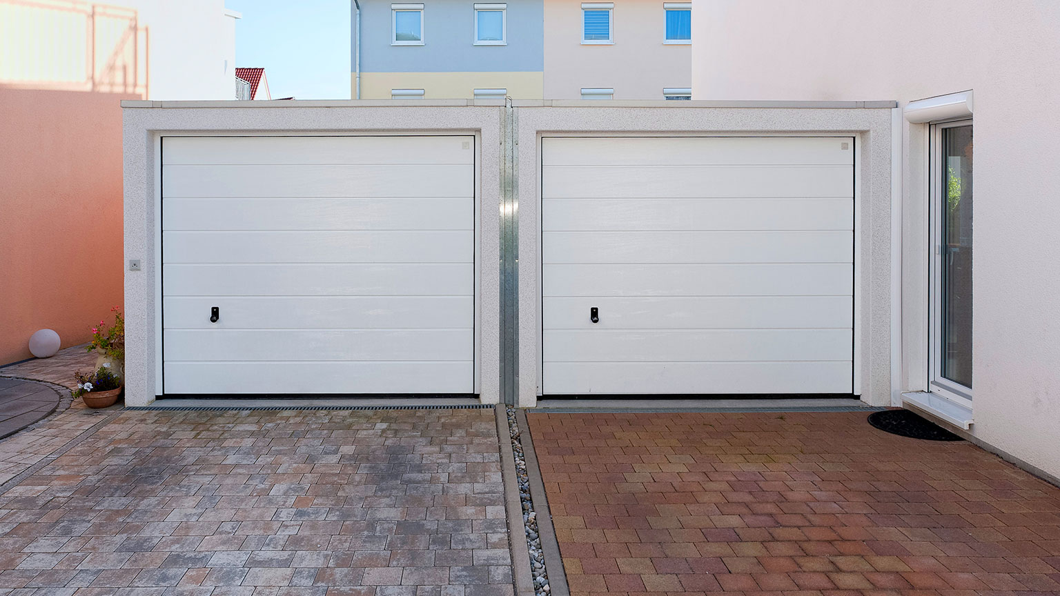 moderne-doppelgarage-beton-mit-zwei-sektionaltoren Moderne Doppelgarage aus Beton mit zwei weißen Sektionaltoren und gepflasterter Einfahrt