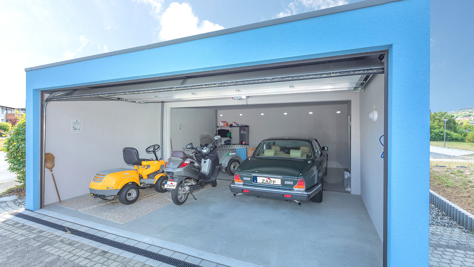 moderne-fertiggarage-innenansicht-mit-auto-und-rasentraktor Innenansicht einer modernen Fertiggarage mit geöffnetem Sektionaltor, Auto, Motorroller und Rasentraktor