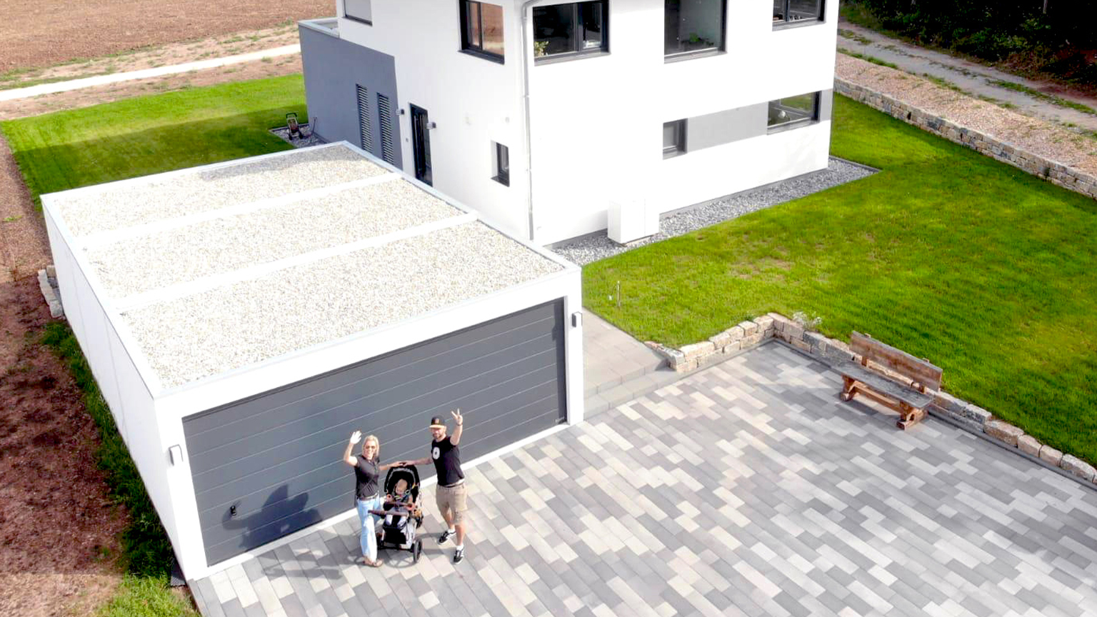 Modern precast concrete garage with flat roof next to a detached house Detached precast concrete garage with extensive green flat roof and anthracite sectional door beside a modern single-family home, paved driveway with family and stroller seen from above, ZAPF quality by ZAPF Garagen