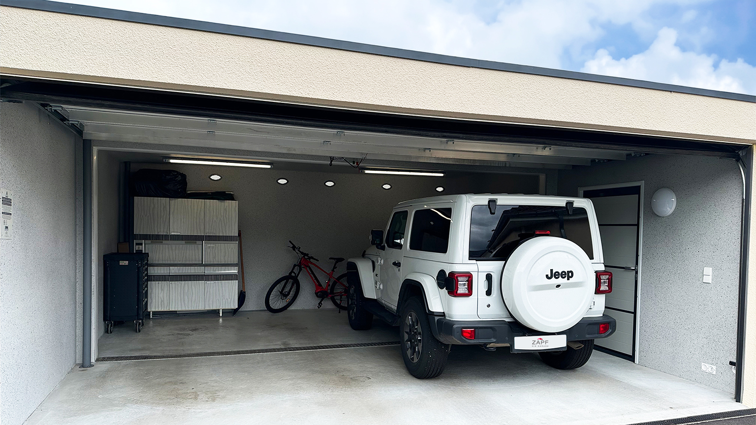 Moderne Einzelgarage mit geöffnetem Sektionaltor und integriertem Stauraum Helle Betonfertiggarage mit geöffnetem Sektionaltor, weißem Geländewagen im Innenraum, seitlichem Zugangstürblatt, Deckenbeleuchtung sowie Schrank- und Regalsystem und Fahrrad im hinteren Bereich, vom Hersteller ZAPF