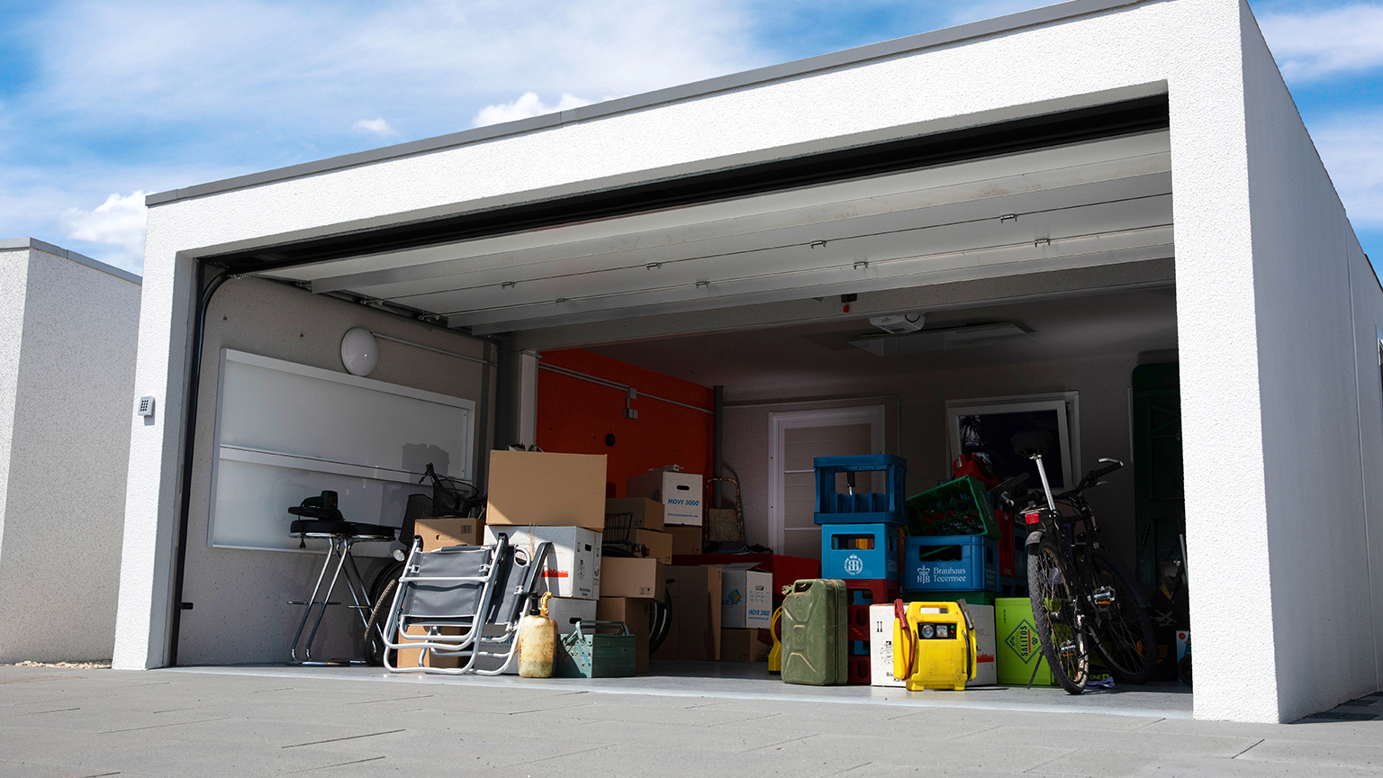 Moderne Fertiggarage mit geöffnetem Garagentor und vielseitigem Stauraum Helle Betonfertiggarage mit geöffnetem Sektionaltor, seitlichem Fenster und Innenraum mit Umzugskartons, Getränkekisten, Fahrrad, Klappstühlen, Kanistern und weiterem Zubehör auf dem Garagenboden, ZAPF Garagen