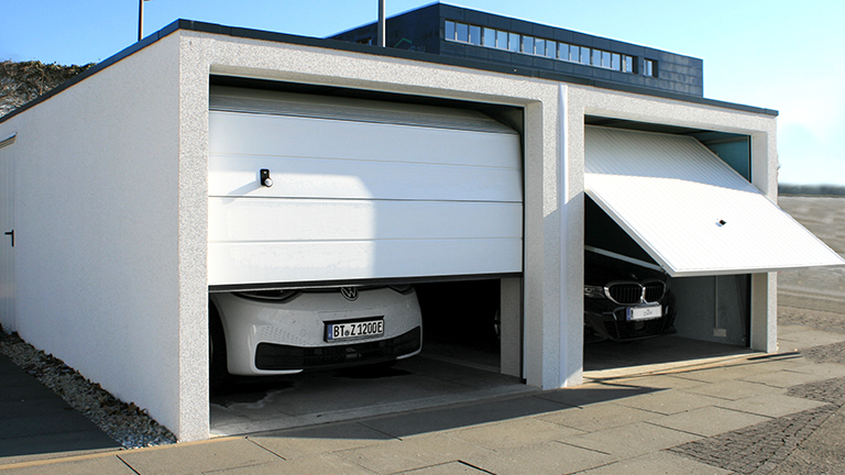 Moderne Betonfertiggarage mit zwei Stellplätzen, links mit geschlossenem weißem Sektionaltor und rechts mit geöffnetem weißem Schwingtor, davor gepflasterte Zufahrt, hochwertige Ausführung in ZAPF Qualität von ZAPF Garagen