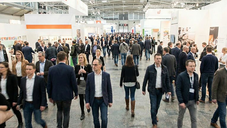 Zahlreiche Besucher bewegen sich zwischen hell gestalteten Messeständen in einer großen Ausstellungshalle mit breiten Gängen, Informationsflächen und Präsentationsbereichen, geschäftiges Treiben auf einer Branchenmesse von ZAPF Garagen
