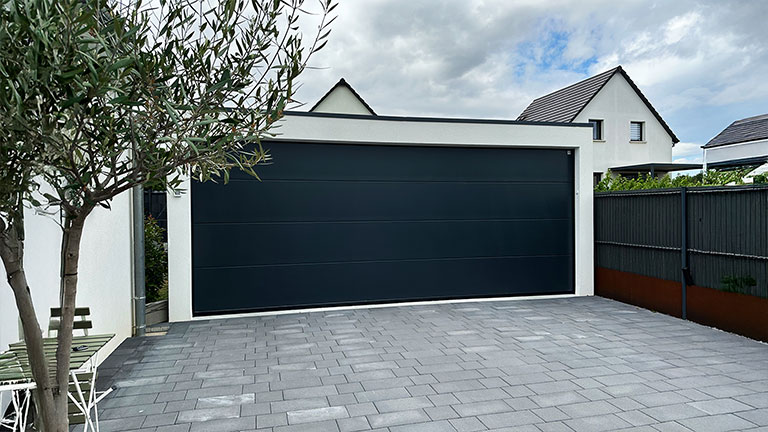 Moderne Großraumgarage mit anthrazitfarbenem Sektionaltor vor weißem Einfamilienhaus, gepflasterte Einfahrt und schwarzer Sichtschutzzaun.
