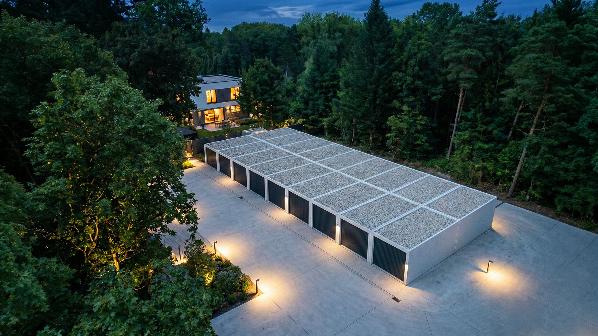 Mehrere aneinandergebaute Betonfertiggaragen mit dunklen Garagentoren und Kiesdach, angeordnet auf großer betonierter Hoffläche mit Beleuchtung, umgeben von dichtem Wald und modernem Wohnhaus im Hintergrund in der Abenddämmerung, Garagenanlage von ZAPF