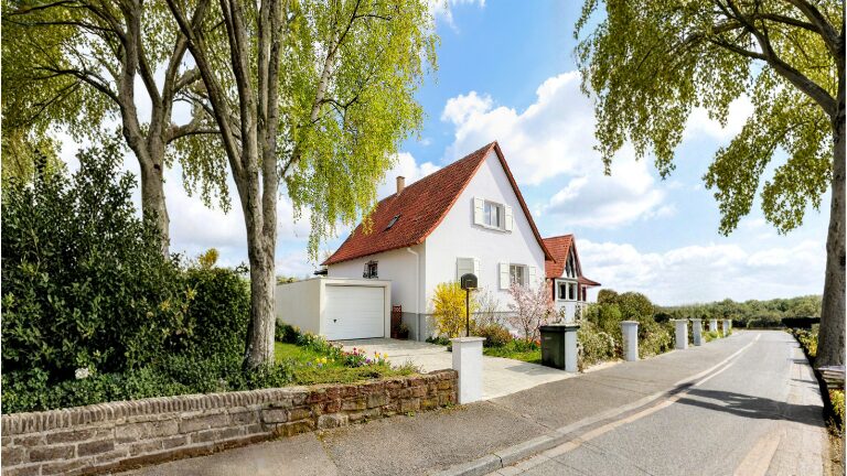 Helles Einfamilienhaus mit rotem Satteldach, integrierter Garage mit weißem Garagentor, gepflasterter Einfahrt, Garten und Bäumen entlang einer ruhigen Wohnstraße, moderne Garagenlösung im Wohnumfeld von ZAPF