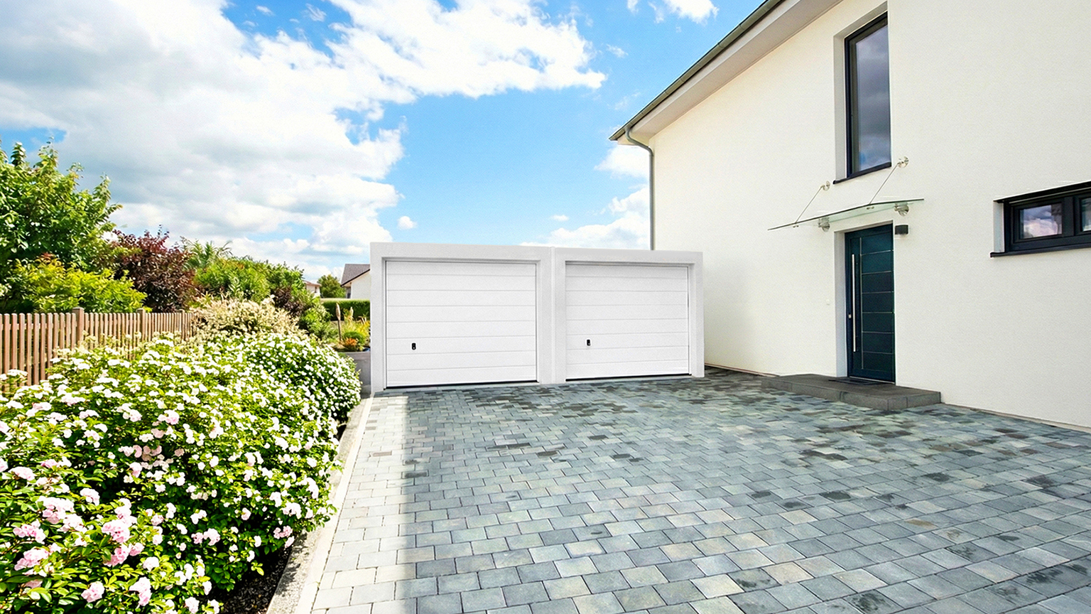 Doppelgarage aus Beton mit zwei weißen Sektionaltoren neben modernem Einfamilienhaus Freistehende Doppelgarage aus Beton mit zwei weißen Sektionaltoren vor einem modernen Einfamilienhaus, große gepflasterte Einfahrt mit Gartenbeeten, Hecke und Blumen entlang eines Holzzauns, ZAPF Garagen