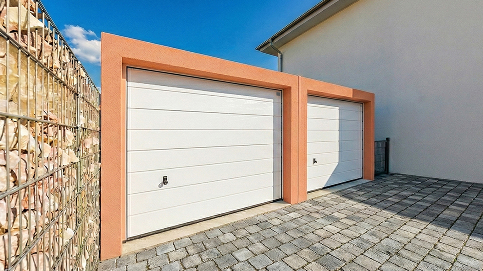 Precast concrete garage with two white sectional doors and orange plaster frame Double precast concrete garage with two white sectional garage doors, orange plastered front frame, paved driveway, gabion wall on the left and house wall on the right under a blue sky by ZAPF