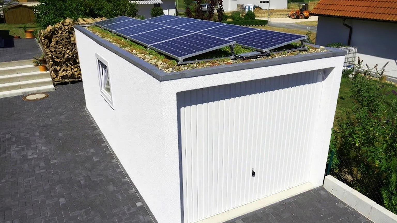 Betonfertiggarage mit Sektionaltor und Photovoltaikanlage auf begrüntem Flachdach Weiße Betonfertiggarage mit vertikal strukturiertem Sektionaltor, kleinem Seitenfenster und Flachdach mit Kies, Dachbegrünung und montierten Photovoltaikmodulen neben gepflasterter Einfahrt und gestapeltem Brennholz von ZAPF