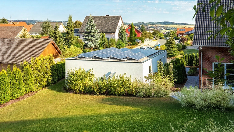 Moderne weiße Betonfertiggarage mit Flachdach und mehreren Photovoltaikmodulen auf der Dachfläche, umgeben von Hecken, Garten und Einfamilienhäusern in einer ruhigen Wohnsiedlung von ZAPF