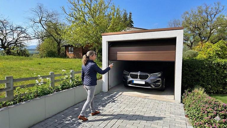Betonfertiggarage mit heller Fassade und geöffnetem braunen Sektionaltor, davor eine Person auf gepflasterter Einfahrt während ein Auto in der Garage steht, umgeben von Garten, Bäumen und Wohnumfeld von ZAPF
