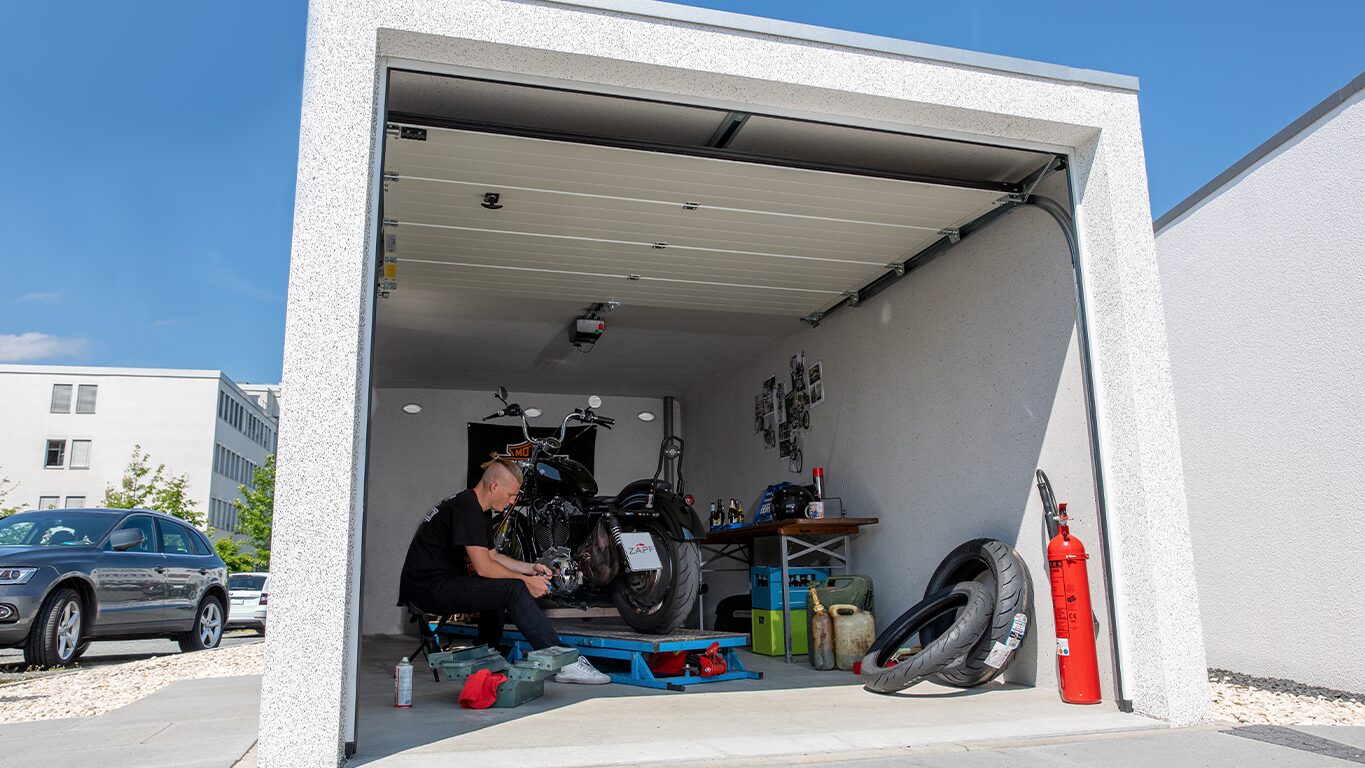 Betonfertiggarage mit geöffnetem Sektionaltor und Motorradwerkstatt Fertiggarage mit geöffnetem Sektionaltor, Person arbeitet an einem Motorrad auf Hebebühne, Werkbank mit Werkzeugen und Kanistern, Reifen und Feuerlöscher im Vordergrund, von ZAPF